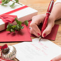 Person writing a letter with Christmas-themed stationery and gifts on a wooden table.