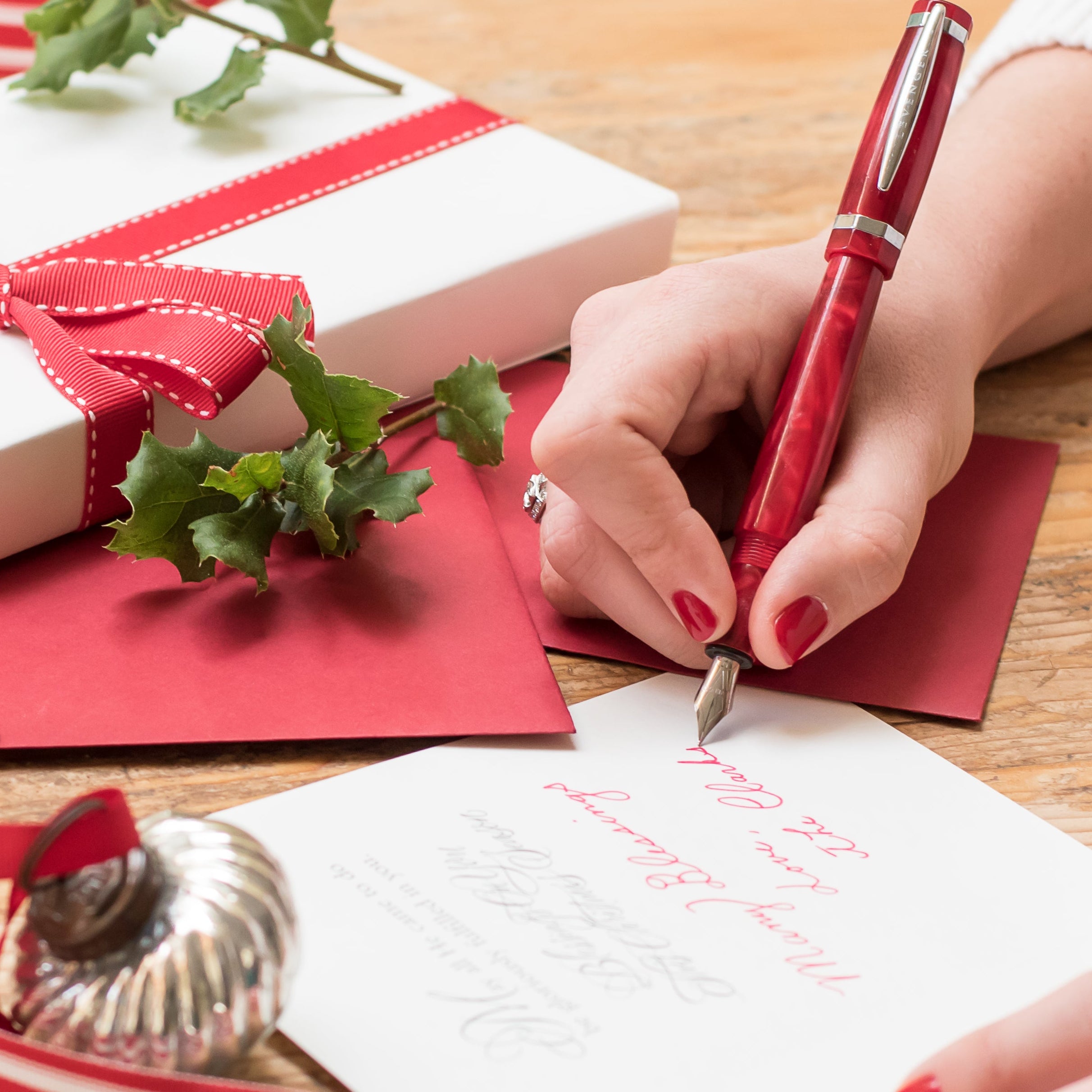 Person writing a letter with Christmas-themed stationery and gifts on a wooden table.